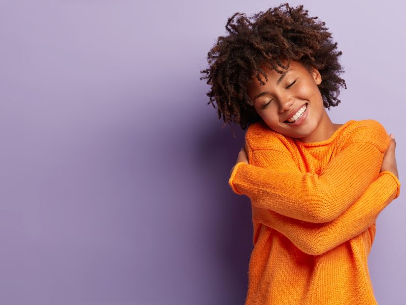 Photo of pleased dark skinned lady with Afro hairstyle, imagines meeting with boyfriend, embraces herself, smiles tenderly, keeps eyes shut, wears orange sweater, isolated over purple background